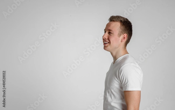 Fototapeta teen guy looking away happy
 and smiling with teeth in the right side of the frame on a white background in a white T-shirt