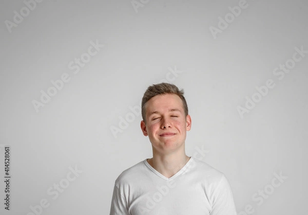 Fototapeta 
teen guy looking at camera closed his eyes happy
 and sincerely smiling with teeth on a white background in a white T-shirt
