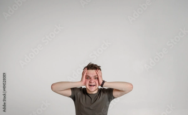 Fototapeta teen guy looking at camera holding his head and can't believe surprised glad
 and sincerely smiling with teeth on a white background in a green T-shirt