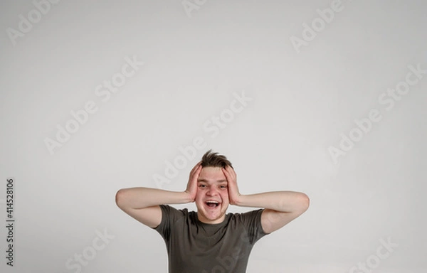 Fototapeta teen guy looking at camera holding his head and can't believe surprised glad
 and sincerely smiling with teeth on a white background in a green T-shirt