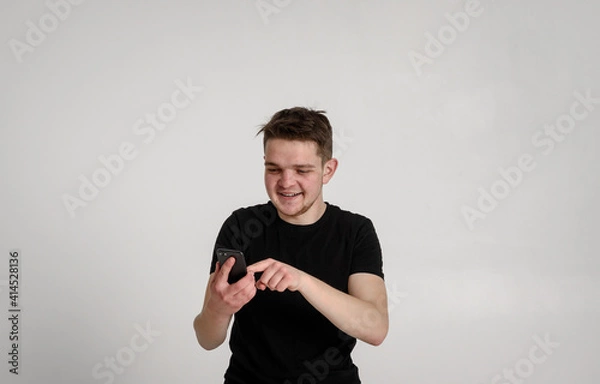 Fototapeta 
teen guy looking at camera
looking for something on the phone searching the internet and smiling sincerely with teeth on a white background in a black t-shirt