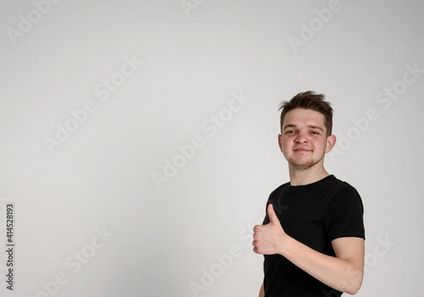 Fototapeta teen guy looks at the camera and shows a like smiling with kind eyes on a white background in a black t-shirt
