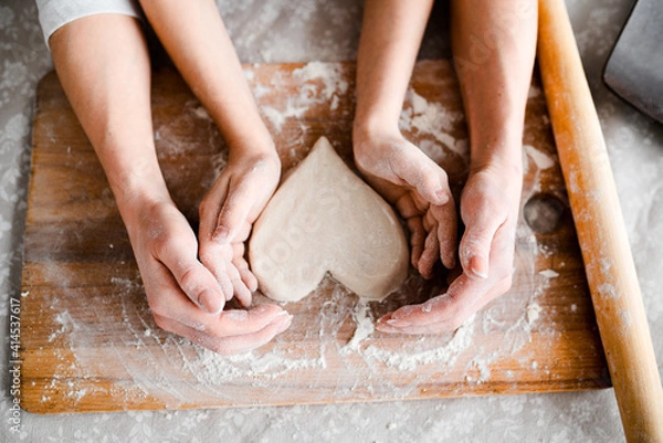 Obraz Top view, hands close-up. Mom and daughter make cookies in the form of Wednesday, together roll out the heart with a rolling pin on a wooden board in flour