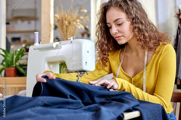Fototapeta young tailor working on sewing machine, hand making clothes in home interior. caucasian curly female sits enjoying process of sewing clothes. creative skills design, hobbies and lifestyle concept