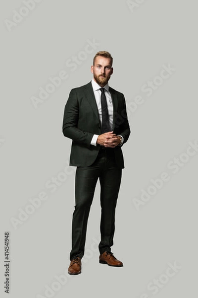Fototapeta Serious looking handsome caucasian businessman looking serious with a beard and wearing a suit and tie, hands clasped on a grey background.