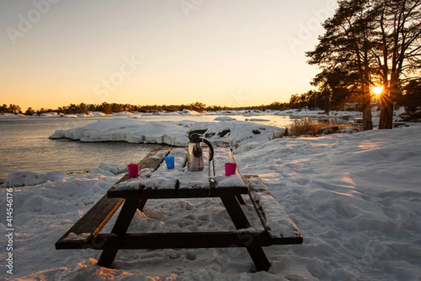 Fototapeta Outdoors coffee break in the winter landscape by the sea during sunset. Thermos and mugs on picnic table covered in snow. Photo taken in the archipelago outside Oskarshamn, Sweden