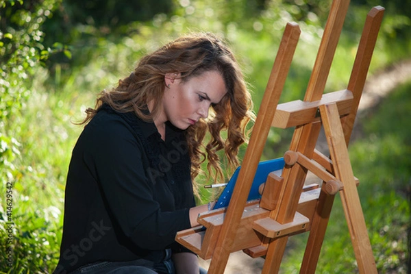 Fototapeta Side view of a portrait of a young white woman drawing a picture on a wooden easel in nature.