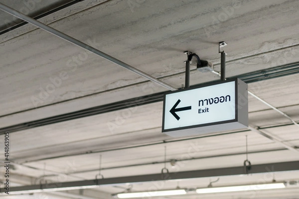 Fototapeta A white building exit sign hung from the ceiling under the lights.