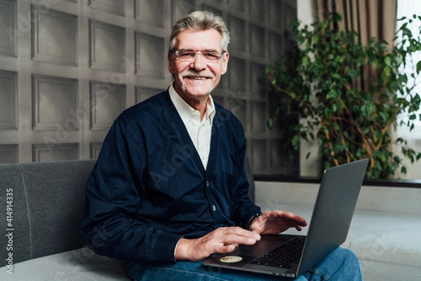 Fototapeta Portrait of a cheerful old grandfather sitting on a sofa with a computer he looks at the camera and smiles