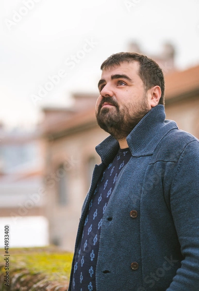 Fototapeta Bearded guy in dark blue sweater leaning on a stone wall in the street, looking at the sky seriously.