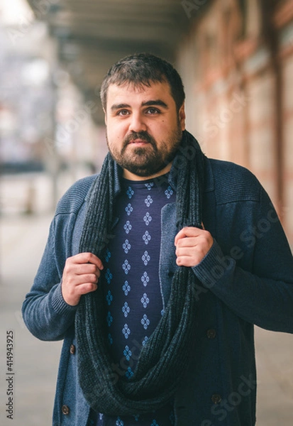 Fototapeta Bearded guy in blue sweater, standing on the street with his hands on his scarf, looking at the camera. 