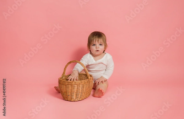 Fototapeta little girl in a white bodysuit sits with a wicker basket on a pink background with space for text