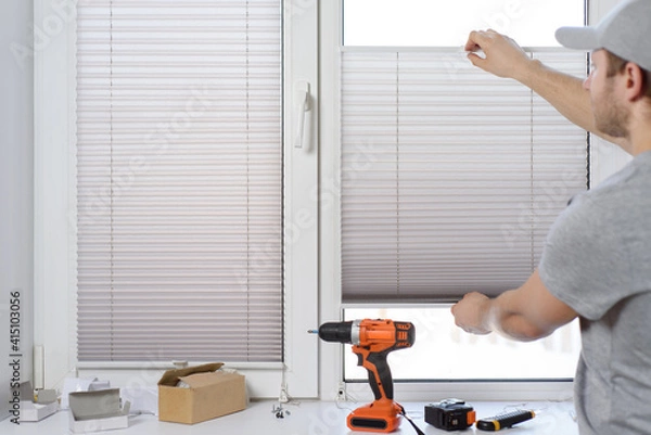 Fototapeta Man installing gray pleated blinds on the window with screwdriver