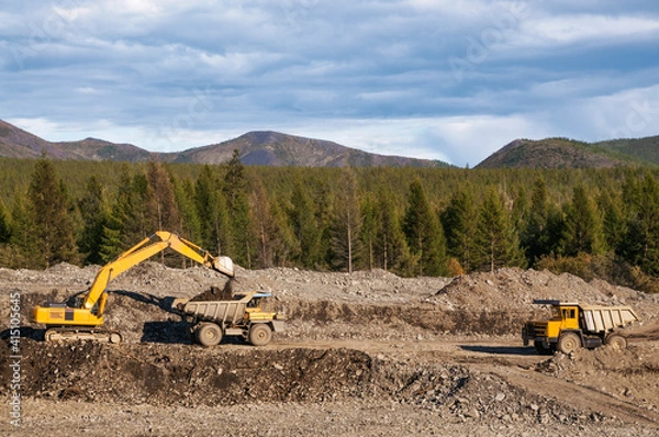 Fototapeta Excavator in the process of work in an industrial mountainous area (loads gold-bearing mountain soil into the body of a mining dump truck). Mining