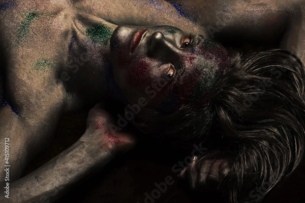 Fototapeta Fashion and art man concept. Guy on mysterious face with fashionable make up, covered with silver paint and colorful glitters. Nude torso covered with shimmering silver paint.