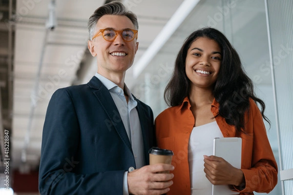 Fototapeta Confident colleagues standing together holding digital tablet and cup of coffee in modern office. Portrait of businesspeople looking at camera, smiling. Successful business concept  