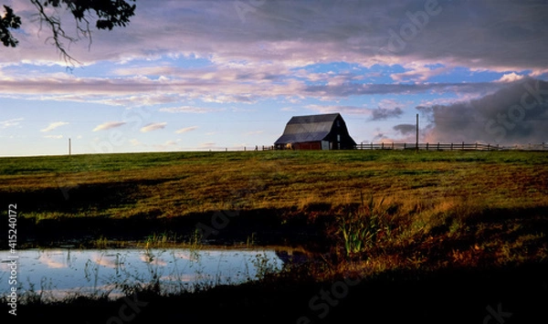 Fototapeta An idyllic scene of a horse in front of a barn at the edge of a pasture with purplish blue/gray clouds in the background and a pond reflecting the clouds in the foreground. Rural Missouri 1990