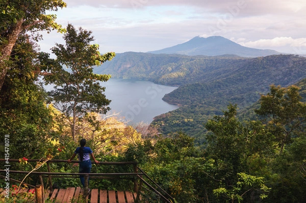 Obraz young woman on a wooden bridge appreciating a beautiful view of the Mombacho volcano and the Apoyo lagoon from sendero los mangos, Catarina, Nicaragua