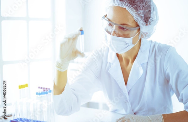 Fototapeta Professional female scientist in protective eyeglasses researching tube with reagents in sunny laboratory toned in blue. Medicine and science researching