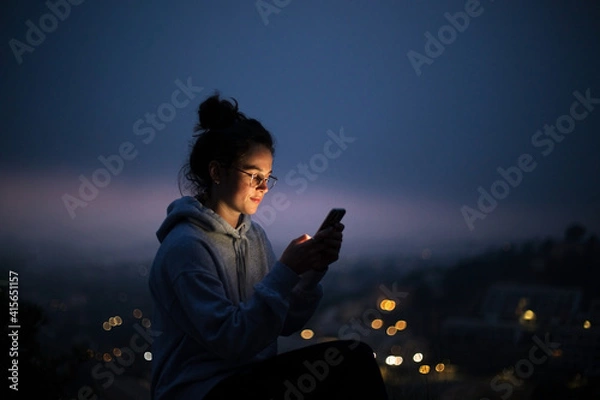 Fototapeta Beautiful young woman use smartphone at night. Millennial generation z female scroll through news feed on app on her phone. Cinematic film look photo of pretty woman at sunset use phone