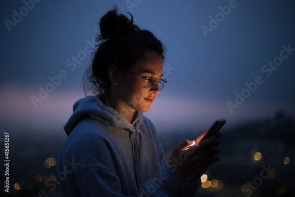 Fototapeta Beautiful young woman use smartphone at night. Millennial generation z female scroll through news feed on app on her phone. Cinematic film look photo of pretty woman at sunset use phone