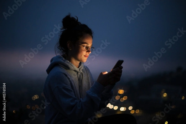 Fototapeta Beautiful and inspiring young woman over epic night city background. Millennial female with screen lit face, cool and trendy teenager looking woman. Smartphone application market, podcast listening