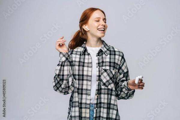 Fototapeta Cheerful young woman wearing wireless earphones dancing while listening popular song on isolated white background, closed eyes. Pretty redhead lady model emotionally showing facial expressions.