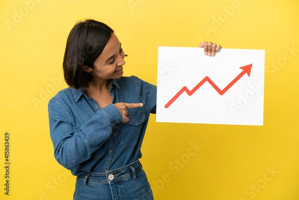 Fototapeta Young mixed race woman isolated on yellow background holding a sign with a growing statistics arrow symbol and  pointing it