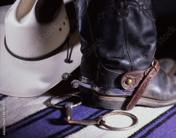 Fototapeta Close up of Black Western Boots with Rowel Spurs, Straw Western Hat and Snaffle Bit with a Purple Wood Pad in Studio Lighting