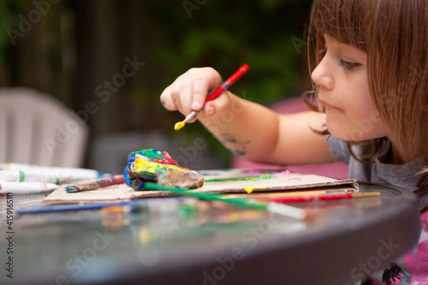 Obraz Young Girl paints Rocks outside on Patio Table to be creative outside on a sunny summer day and surrounded by nature
