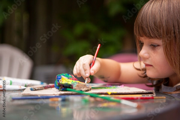 Obraz Young Girl paints Rocks with colorful Paints and Brushes outside to be creative outside on a sunny summer day and surrounded by nature