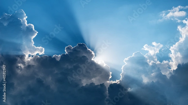 Obraz Sun behind dark clouds with sun rays on blue sky background