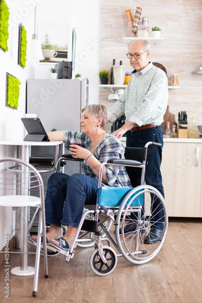 Fototapeta Disabled senior woman in wheelchair using tablet computer in kitchen with husband near. Paralysied handicapped old elderly person using modern communication online internet web techonolgy.