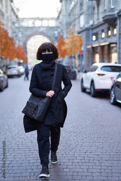 Obraz Pretty trendy young woman in a stylish total black style with a black eco bag enjoys a walk along the street. Attractive girl in fashionable clothes in the European city background