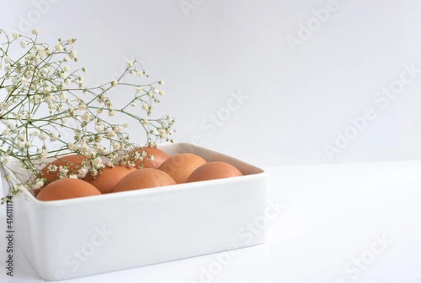 Obraz Easter eggs in a white square dish, on a white table, with a sprig of delicate flowers. Close-up, space for text