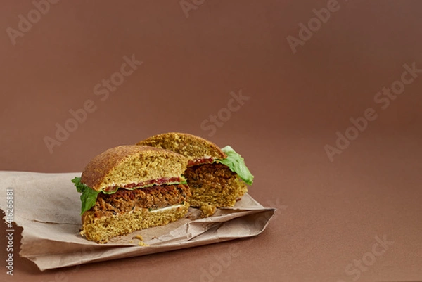 Fototapeta Vegetarian burger in the cut made with quinoa, chick peas, rolled oats, crushed onions and garlic, spices on a Whole Wheat Bun with salat, tomatoes and cucumber on the paper bag. On the left side