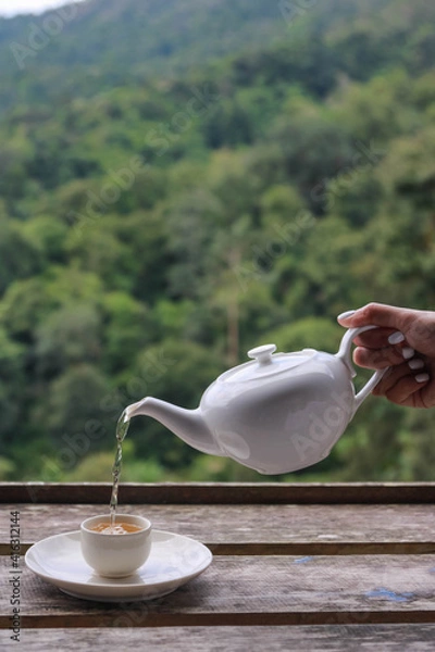 Fototapeta A Woman hand pouring a tea into the cup on the wooden table which has the background of mountain and forest