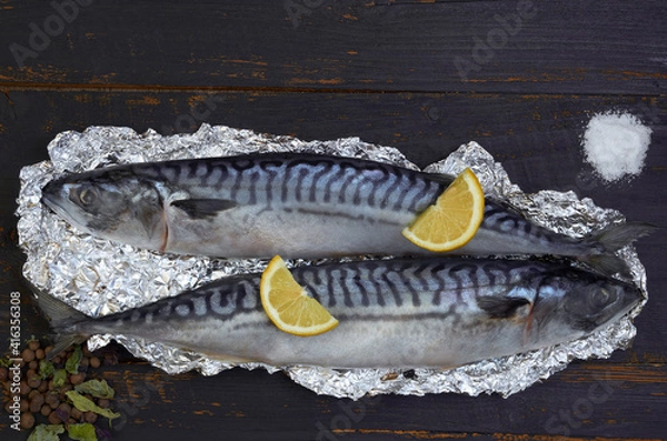 Fototapeta Two atlantic mackerel fishes ready for cooking decorated with spices, salt and lemon slices on the black wooden background. Top view