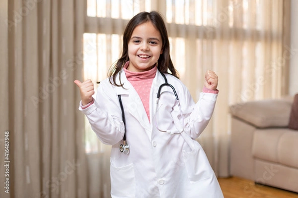 Fototapeta Little child dressed in doctor's uniform giving thumbs up in a fun and positive attitude. She is at home. Space for text.