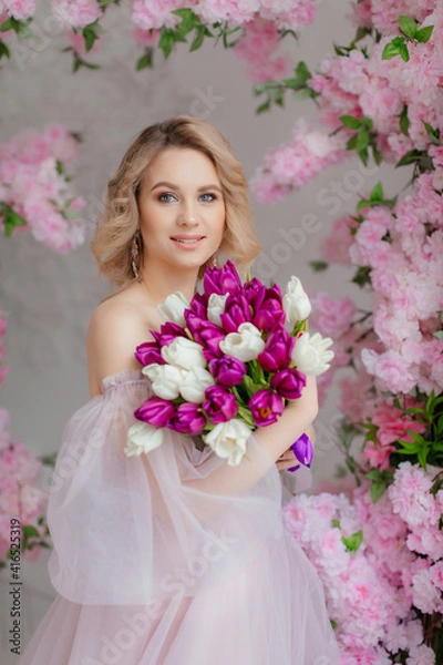Fototapeta Beautiful girl in a lush pink dress in a photo studio. Bouquet of tulips. Blonde girl with tulips. An attractive young woman holds a bouquet of white and purple tulips in her hands.