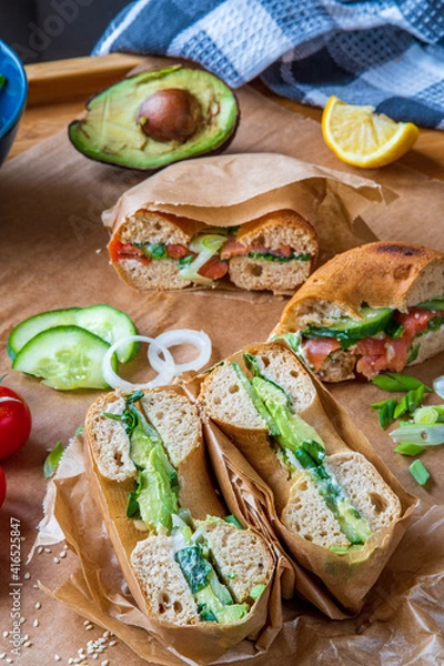 Obraz Stuffed toasted bagel sandwich cut into half with avocado, mayo, cucumber, lettuce and onions. Salmon bagel in the background, wrapped with paper. Gluten free healthy breakfast, vibrant fun colors