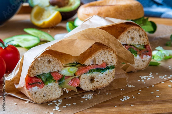 Obraz Stuffed toasted bagel sandwich cut into half with smoked salmon, cream cheese, cucumber lettuce. Avocado bagel in the background on a baking sheet. Gluten free healthy breakfast, vibrant fun colors 