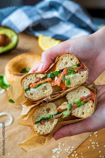 Fototapeta Hands holding stuffed toasted bagel sandwich cut into half with smoked salmon, cream cheese, cucumber lettuce. Avocado bagel in the background. Gluten free healthy breakfast, vibrant fun colors