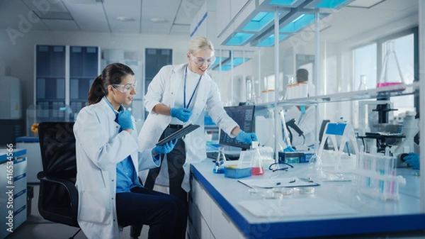 Fototapeta Modern Medical Research Laboratory: Two Female Scientists Working Using Digital Tablet, Analysing Biochemicals Samples. Scientific Lab for Medicine, Microbiology Development. Advanced Equipment