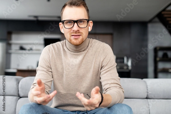 Obraz Portrait of intelligent young bearded hipster man looking at camera, wearing glasses and wireless earphones, sitting on a couch in the living room and gesturing. The view while video-calling concept