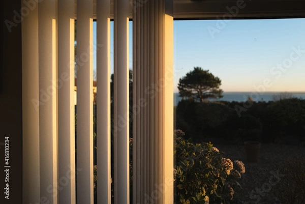 Fototapeta Half open vertical blinds with a golden glow as the sun sets.There is a tree and a view of the sea through the window.
