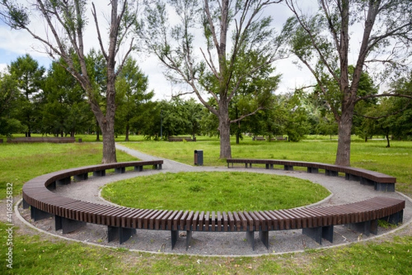 Fototapeta Round wooden bench in the park, standing under the trees. Modern fashionable design round circular bench in the garden.