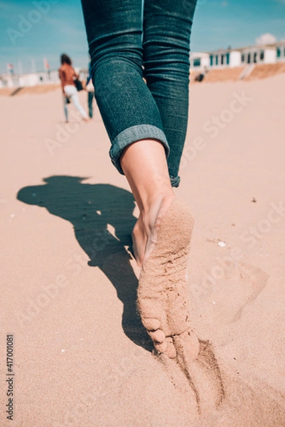 Fototapeta Beach travel - woman in a jeanse walking on sand beach leaving footprints in the sand. Closeup detail of female feet and sand on Holland coast, North Sea