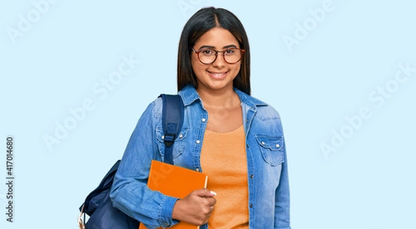Fototapeta Young latin girl wearing student backpack and holding books with a happy and cool smile on face. lucky person.