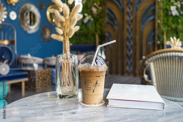 Obraz Iced coffee with straw on the white marble table with book and glass vase of dried decoration plant.